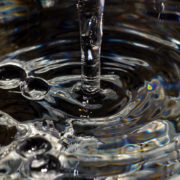 Das querformatige Foto zeigt in Nahaufnahme wie sich ein Wasserstrahl in ein mit Wasser gefülltes Gefäß ergießt.