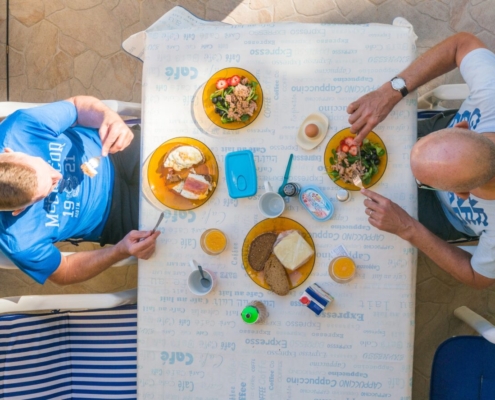 Das querformatige Foto zeigt zwei Männer beim Frühstück auf einem Balkon.