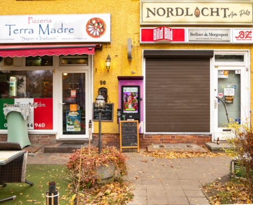 Das querformatige Foto zeigt eine kleine Ladenzeile mit zwei geschlossenen Geschäften: einer Pizzeria und einem Zeitungsladen.