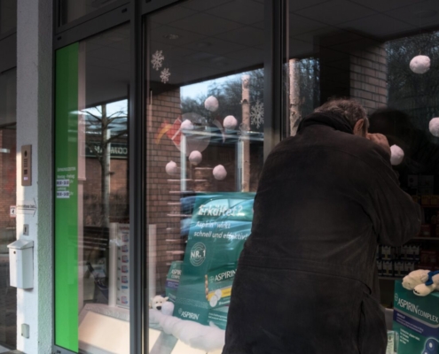 Das querformatige Foto zeigt den Rücken eines Mannes, der durch die Schaufensterscheibe in das Inner einer Apotheke schaut.