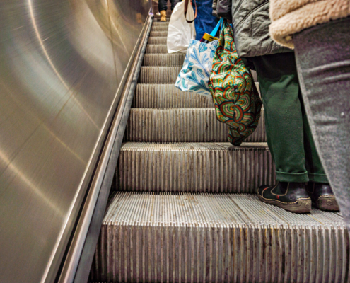 Das querformatige Foto zeigt eine Rolltreppe, auf der Menschen mit vollen Einkaufsbeuteln stehen